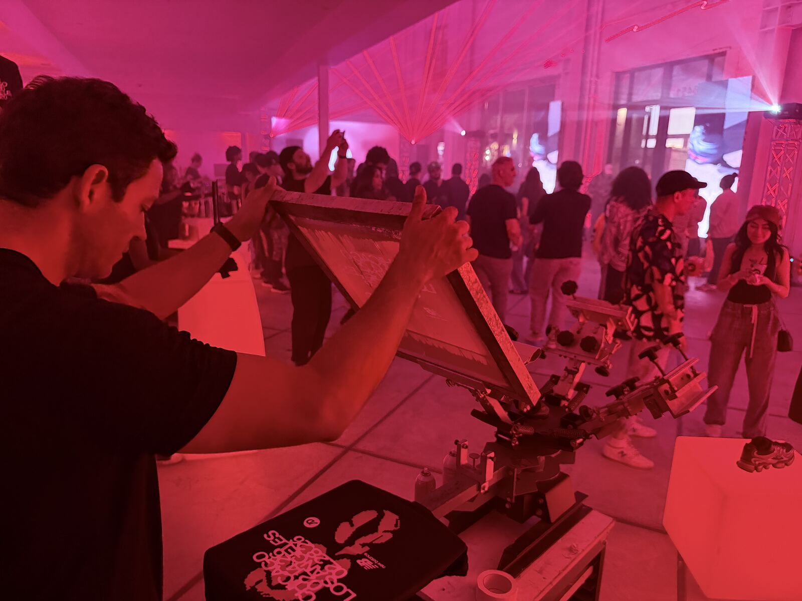 Busy event booth with multiple attendees interacting with a live printing demonstration under vibrant magenta and pink lighting